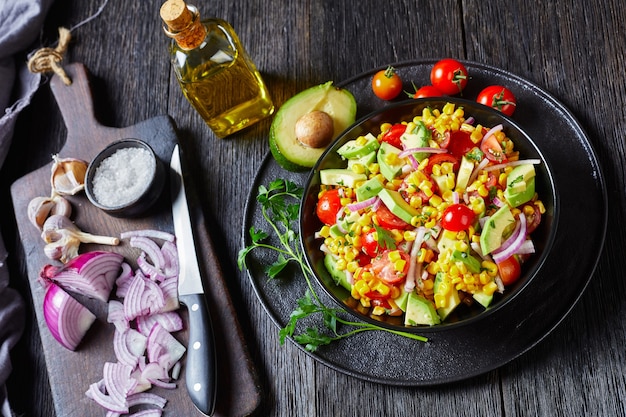 Sweet corn salad with avocado, red onion slices and tomatoes in a black bowl on a dark wooden table with ingredients on dark old cutting board, mexican cuisine, horizontal view from above