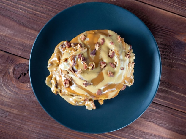 Sweet delicious dessert a cinnamon bun sprinkled with pecans and poured with caramel topping on a black plate and a turd background Top view flat lay