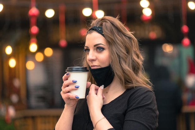 A sweet female of caucasian appearance in a black face mask, sits and works in an Internet cafe