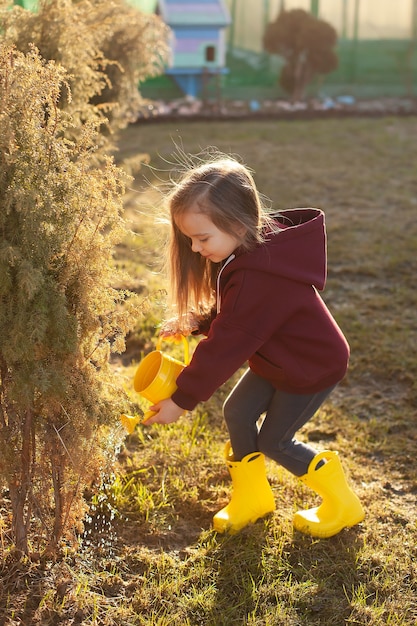 A sweet girl in the spring in yellow boots watering bushes in the garden