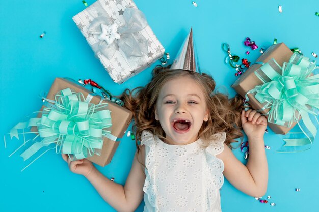 A sweet happy little girl in a cotton white dress on a blue background in a festive hat with gifts and confetti shouts hurrah and laughs with joy celebrating her birthday a place for text