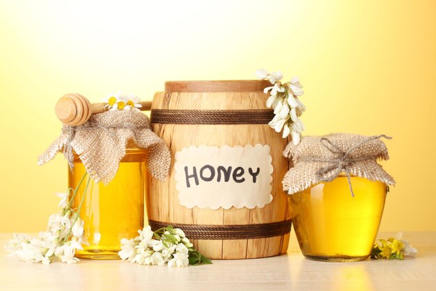 Photo sweet honey in barrel and jars with acacia flowers on wooden table on yellow background