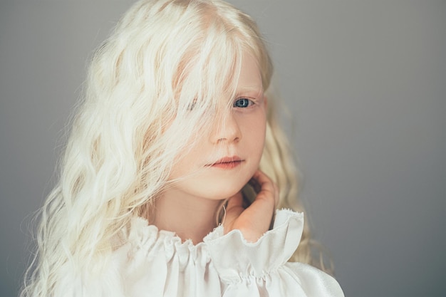 Sweet kid portrait. Natural beauty. Carefree childhood. Cute happy shy albino blonde little angel girl with curly hair blue eyes in white vintage blouse isolated on gray background.