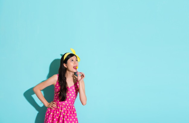 sweet lovely lady eating candy lollipop looking at empty area daydreaming in blue background.