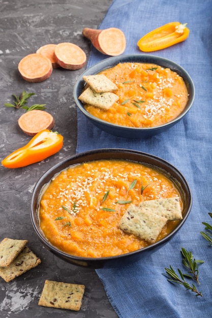 Sweet potato or batata cream soup with sesame seeds and snacks in blue ceramic bowls on a black concrete surface with blue linen textile