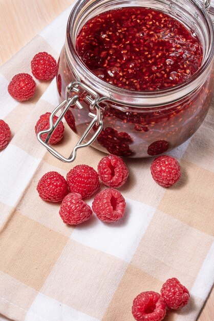 Sweet raspberry jam in a jar and scattered raspberries on a check napkin