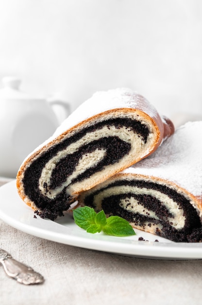 Sweet roll with poppy seeds from yeast dough sprinkled with icing sugar Cut in half Lies on a white plate Decorated with a sprig of mint Home made In the background is a white porcelain teapot