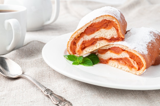 Sweet roll with yeast dough apple sprinkled with icing sugar Cut in half Lies on a white plate Decorated with a sprig of mint Home made In the background is a white porcelain teapot