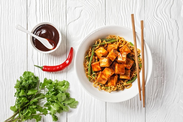 Sweet and spicy asian noodle with hoisin baked crunchy tofu and  green bean  in a white bowl with chopsticks horizontal view from above flat lay