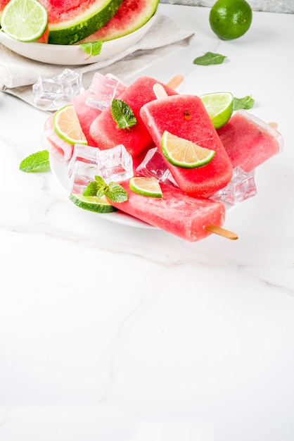 Sweet summer watermelon and lime popsicles with sliced watermelon and mint, on white marble surface 