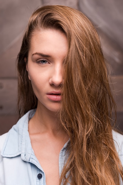 Sweet temptation. Portrait of a beautiful young woman in shirt covering half of her face by hair and looking at camera while standing against metal background