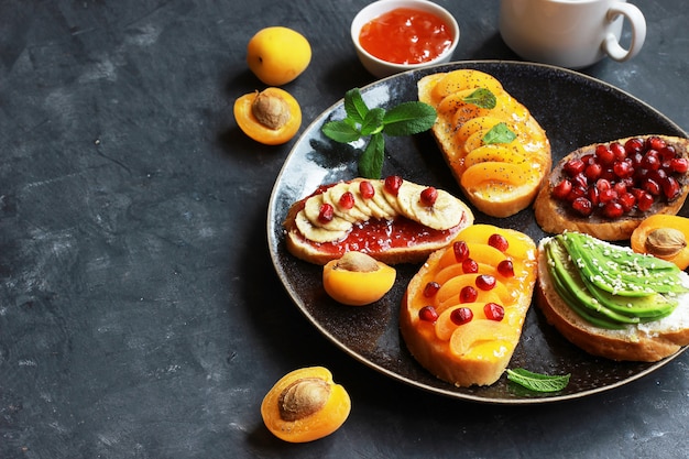 Sweet toast with jam and various fruits apricots,bananas,pomegranates and avocado on the dark background copy space top view.