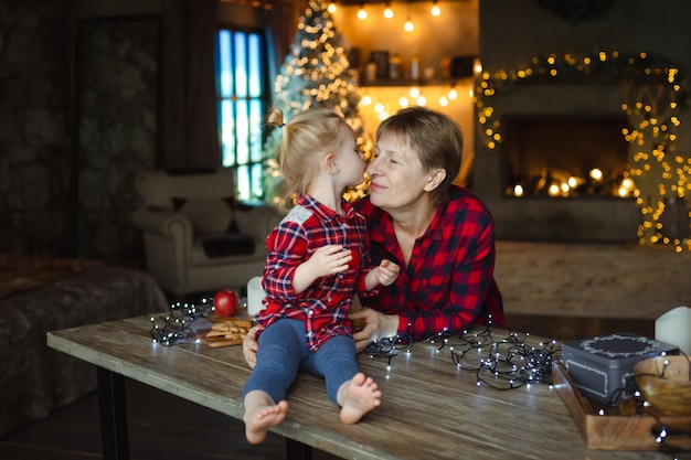 A sweet toddler kisses her grandmother in the nose, sitting on a wooden table in a hunting house decorated for Christmas.