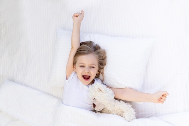 A sweetly yawning baby girl sleeps on a bed on a white cotton pillow under a blanket hugs a teddy bear toy healthy baby sleep at night