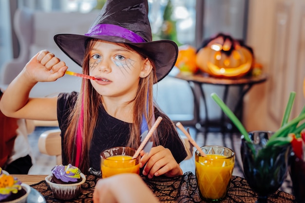 Sweets and juice. Cute beautiful girl wearing wizard Halloween suit enjoying thematic sweets and orange juice