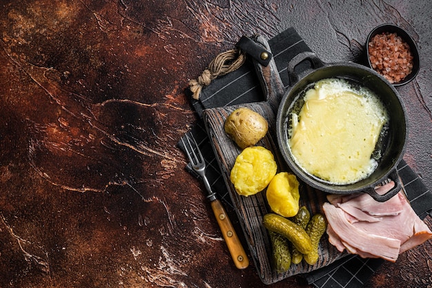 Swiss Raclette Melted cheese with boiled potato and ham on wooden board Dark background Top view Copy space