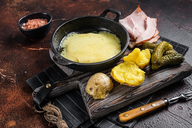 Swiss Raclette Melted cheese with boiled potato and ham on wooden board. Dark background. Top view.