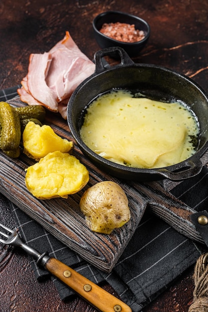 Swiss Raclette Melted cheese with boiled potato and ham on wooden board Dark background Top view