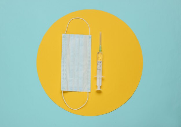 Syringe and medical mask on a blue with yellow circle in the middle