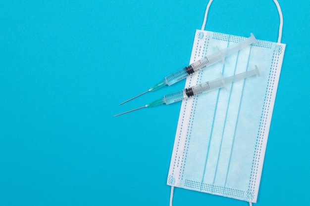 Syringe with medical mask on blue table flat lay