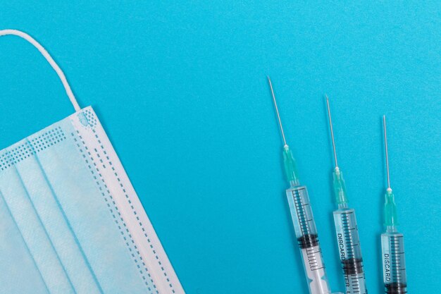 Syringe with medical mask on blue table flat lay