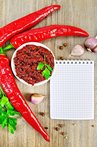 Tabasco in a white cup, fresh red peppers, garlic, peppercorns, parsley, notepad on an old wooden board