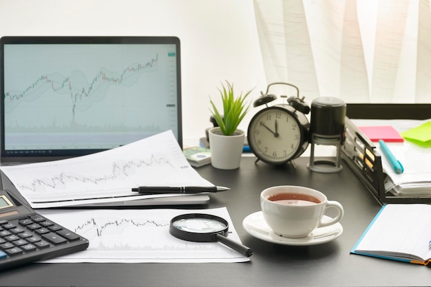 On table are charts of stock price movement there is cup of tea calculator laptop paper for notes