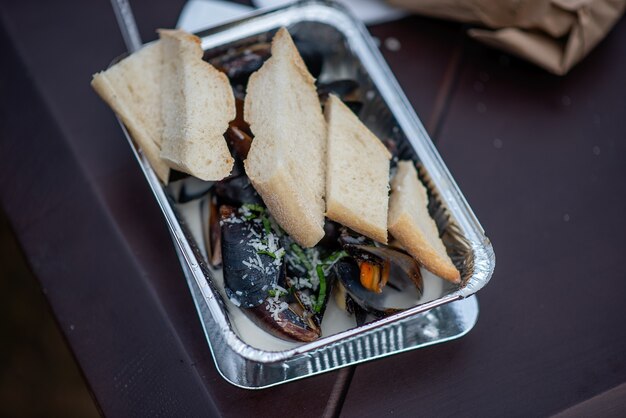 On the table is a disposable foil plate containing fire-baked mussels in garlic sauce, sprinkled with parmesan cheese and a crispy fresh baguette. Street takeout fast food at the festival. Copy space