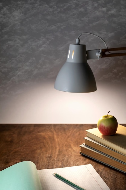 Table lamp illuminates the workplace on the office desk a stack of books an open notebook a pencil and an apple on a gray background with clouds