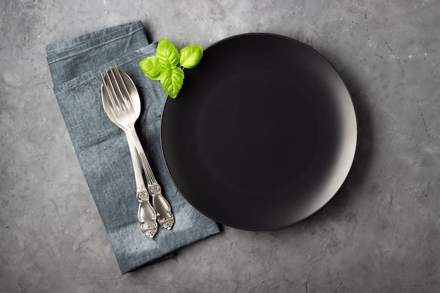 Table setting. Black plate, cutlery, napkin and basil leaves over gray concrete