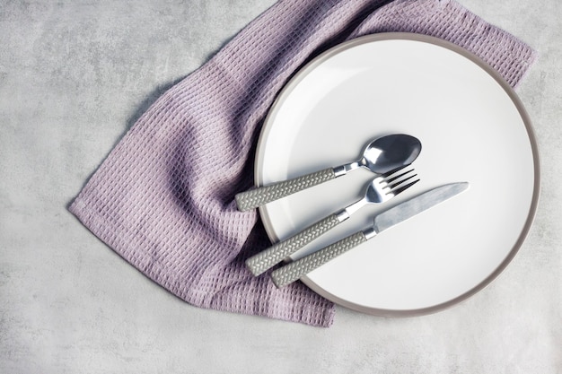 Table setting. White plate, cutlery and napkin on light gray background. Flat lay, top view.