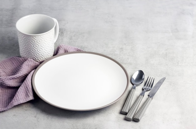 Table setting. White plate, mug, cutlery and napkin on light gray background.