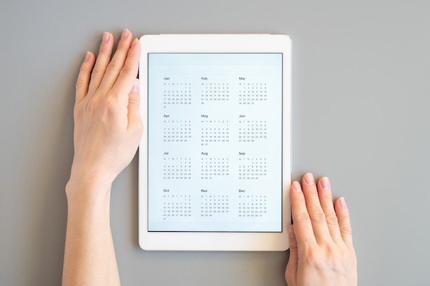 Tablet computer with an open app of calendar for unspecified unknown date year without date in a womans hands on a gray background concept business or to do list goals top view flat lay