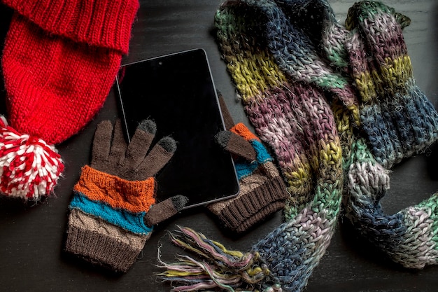 The tablet is on the gloves, as if lying in the hands of the person near to a black wooden table are other colorful winter clothes: a red hat and scarf