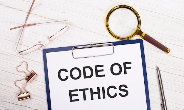 A tablet with the text CODE OF ETHICS on white paper a magnifier glasses golden paper clips and a pen lie on a white office table Top view flat lay