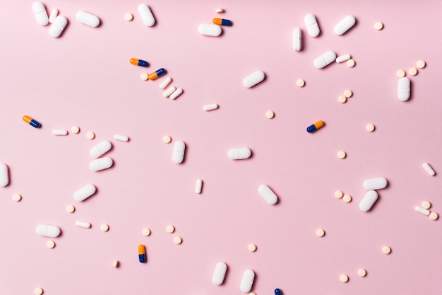 Tablets and capsules of medicines of different colors and sizes randomly scattered on a pink surface