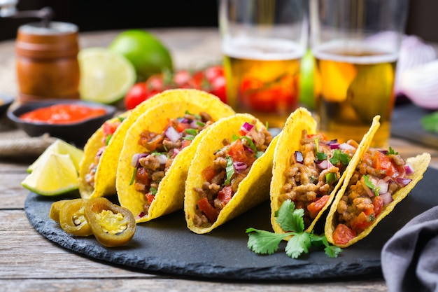 Photo taco shell ellow corn tortilla chips nachos with ground beef, mince, guacamole, red hot jalapeno chili salsa and cheese sauce with tequila or beer on a table.