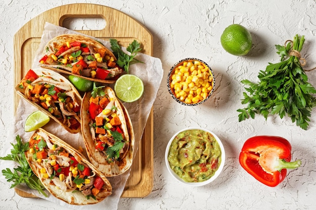 tacos with grilled chicken meat, corn, roasted sweet potatoes cubes, red pepper  and parsley served  on a wooden board on  a white concrete table