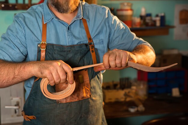 Photo tailor in apron working with leather at workshop, belt manufacturing process