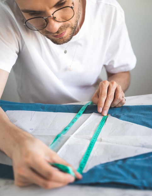 A tailor uses a measuring tape during his work in home workshop A selftaught seamster in white tshirt and glasses works with fabric pattern and chalk Hobby small business and life style concept