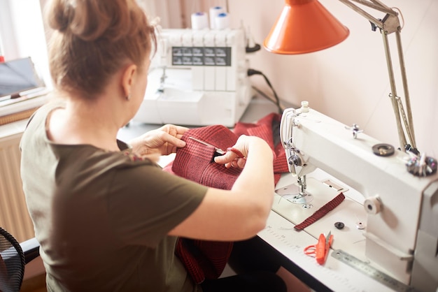 Tailor woman sitting on workplace in atelier and using scissors for cutting off cloth piece in sewing process Back view