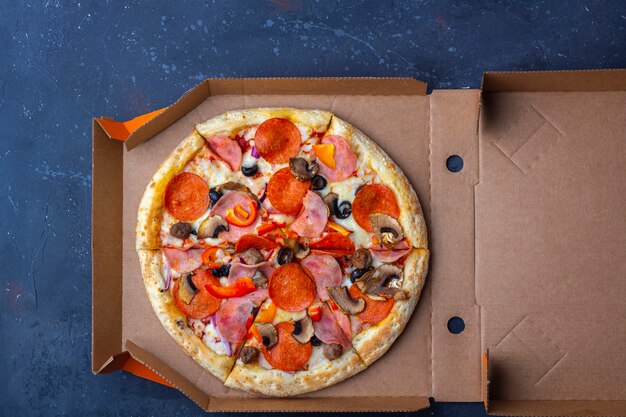 Takeaway delivery service. Opened cardboard box with fresh prepared pizza with mushrooms, ham and cheese on a dark background. Fast food concept.