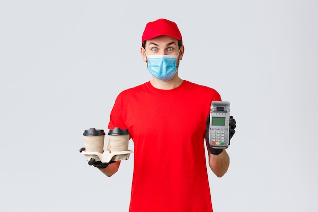 Takeaway, food and groceries delivery, covid-19 contactless orders concept. Excited and surprised client or employee in red uniform, face mask and gloves, giving client paying terminal with coffee