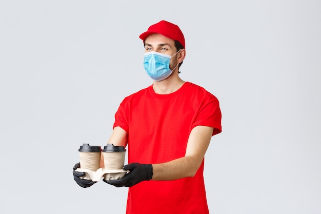 Takeaway, food and groceries delivery, covid-19 contactless orders concept. Pleasant courier in red uniform, face mask and gloves, giving out coffee to customer, stand grey background