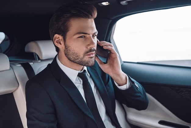 Taking everything seriously. Handsome young man in full suit talking on the smart phone while sitting in the car