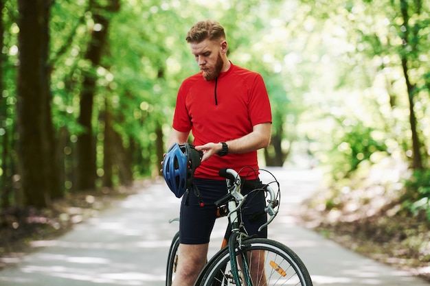 Taking off protective helmet. Cyclist on a bike is on the asphalt road in the forest at sunny day