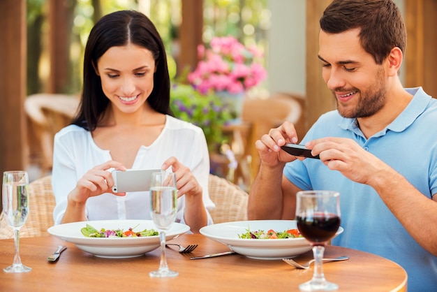 Taking pictures of food. Beautiful young loving couple taking pictures of their food and smiling while relaxing in outdoors restaurant together