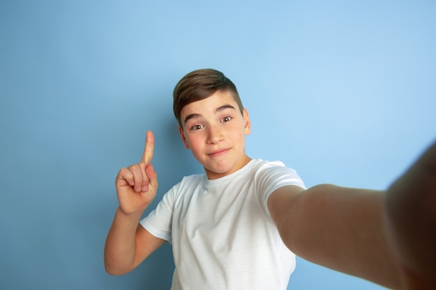 Taking selfie with pointing up. Caucasian boy portrait isolated on blue studio background. Beautiful teen male model in white shirt posing. Concept of human emotions, facial expression, sales, ad.