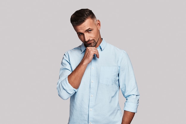 Taking time to think. Thoughtful young man keeping hand on chin and making a face while standing against grey background