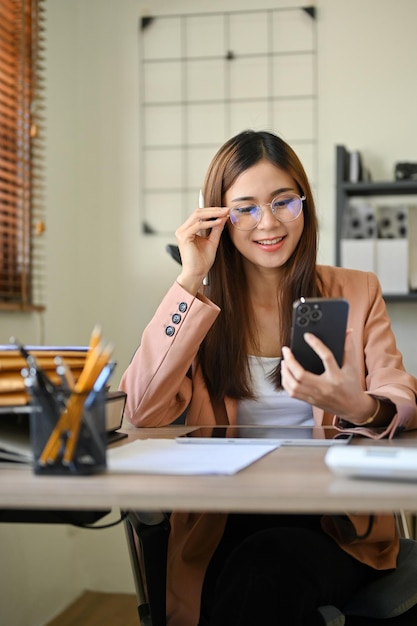 Talented Asian businesswoman in eyeglasses using her mobile phone at her office desk